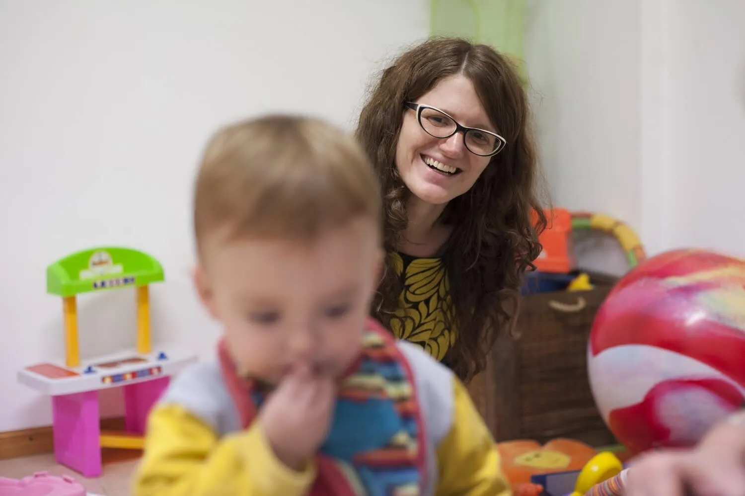 A caregiver smiling as a toddler explores toys